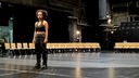 Auf dem Foto ist eine schwarze Frau, die auf eine Theaterbühne steht. Hinter ihr sind leere Stühle.