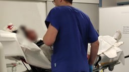 Auf dem Foto ist ein Mann mit Brille in blauer Kleidung, der zu einem Patienten auf einer Liege spricht. Der Kopfverband des Patienten ist voller Blut.