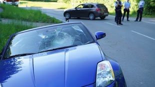 Frontalansicht: Verunfalltes blaues Auto liegt im Straßengraben mit gesprungener Scheibe. Im Hintergrund Polizei und weiteres Auto
