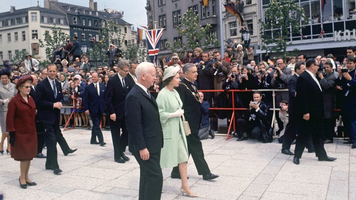 Königin Elisabeth II. und Bundespräsident Lübke gehen mit anderen Personen durch eine mit Absperrgittern geschützte Straße