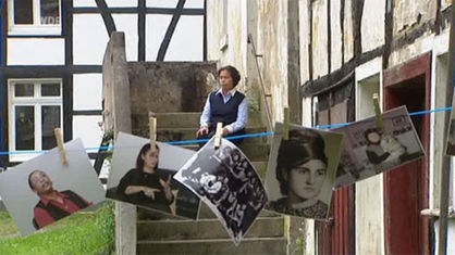 Eine ehemalige Gastarbeiterin sitzt auf einer Treppe, vor ihr hängen auf einer Wäscheleine Bilder von weiteren Gastarbeiterinnen, die annodazumal nach Deutschland gekommen waren.