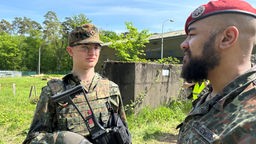 Zwei Soldaten stehen in der Natur und schauen sich an, der eine hat ein Gewehr in der Hand.