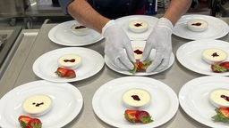 Mehrere Dessertteller mit einem weißen Mousse stehen auf einer silbernen Arbeitsfläche, zwei Hände in Einmalhandschuhen legen Erdbeeren auf einen der Teller