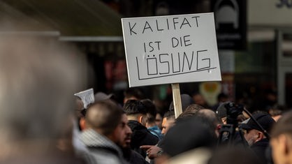 Teilnehmer einer Islamisten-Demo halten ein Plakat mit der Aufschrift "Kalifat ist die Lösung" in die Höhe. Foto vom 27.04.2024